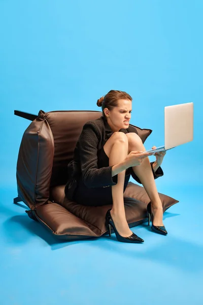 商业女性坐在倒着的扶手椅上 看着笔记本电脑 脸上带着不愉快的表情和蓝色的工作室背景作对比 业务概念 日常工作 截止日期 自由应聘人员 办公室 — 图库照片