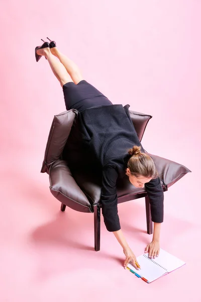 奇妙なことに アームチェアに横たわる女性は 作業計画とノートを見て ピンクのスタジオのバックグラウンドで床に取り組んでいます ビジネス ワーキングルーチン 締め切り フリーランス オフィス — ストック写真