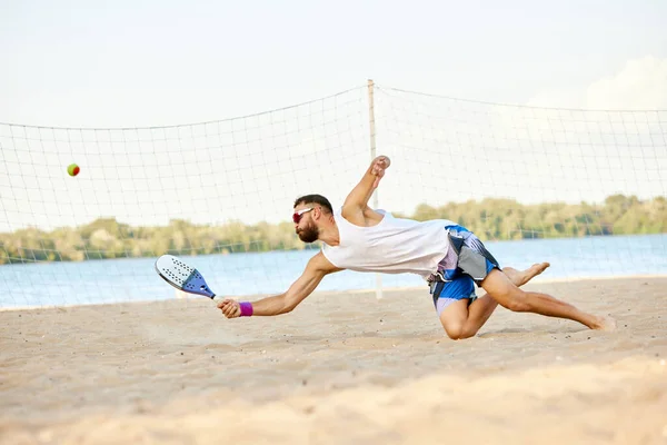 Dynamic image of young man playing beach, paddle tennis, hitting ball with racket and falling down. Outdoor training on warm summer day. Concept of sport, leisure time, active lifestyle, hobby, ad