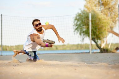 Kumsalda oynayan, raket oynayan, raketle top oynayan ve yere düşen genç bir adam. Sıcak bir yaz gününde açık hava eğitimi. Spor anlayışı, boş zaman, aktif yaşam tarzı, hobi, oyun, reklam