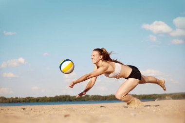 Plaj voleybolu oynayan rekabetçi genç bayan sporcu, elle topa vuruyor ve kuma düşüyor. Eşleşme. Spor anlayışı, aktif ve sağlıklı yaşam tarzı, hobi, yaz zamanı, reklam