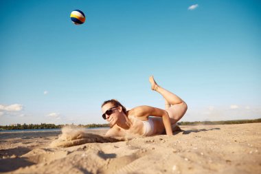 Hareket halindeki genç bir kadının dinamik görüntüsü, plaj voleybolu oynarken, topa vurup kumun üzerine düşerken. Spor anlayışı, aktif ve sağlıklı yaşam tarzı, hobi, yaz zamanı, reklam