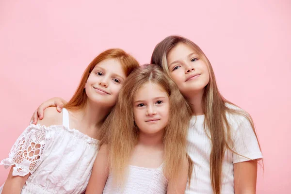 stock image Three beautiful little girls, children in white clothes standing closely to each other, looking at camera against pink studio background. Concept of skincare, childhood, health, beauty products, ad