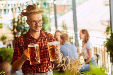 Kareli gömlekli ve fötr şapkalı bira içen genç adamın yakın çekimi. Arka planda bulanık insanlar. Güneşli bir gün. Oktoberfest kavramı, geleneksel zevk, arkadaşlık, boş zaman, keyif