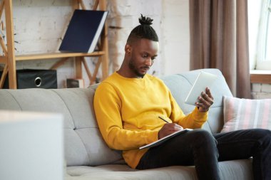 Genç Afrikalı bir adam, evdeki kanepede oturan bir öğrenci, tablet dersleri izliyor, notlar alıyor. Online üniversite. İş ve eğitim kavramı, serbest meslek, modern yaşam tarzı