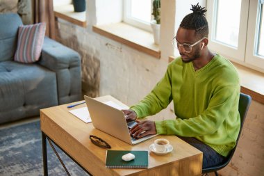 Genç Afrikalı adam evde masada oturuyor, dikkatle ve dikkatle dizüstü bilgisayarla çalışıyor. Çevrimiçi projeler, uzaktan çalışma. İş ve eğitim kavramı, serbest meslek, modern yaşam tarzı