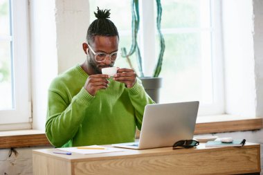 Yoğunlaşmış, genç, Afrikalı bir adam evde oturmuş, kahve içiyor, dizüstü bilgisayara bakıyor ve uzaktan çalışıyor. İş ve eğitim kavramı, serbest meslek, modern yaşam tarzı