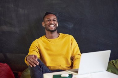 Genç Afrikalı bir adam dizüstü bilgisayarda uzaktan çalışıyor, gülümsüyor, iş arkadaşlarıyla çevrimiçi video görüşmeleri yapıyor. İş ve eğitim kavramı, serbest meslek, modern yaşam tarzı