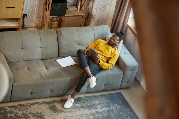 Top view image of young african man in yellow sweater, sitting on sofa in living room at home and watching videos on tablet. Concept of business and education, freelance job, modern lifestyle