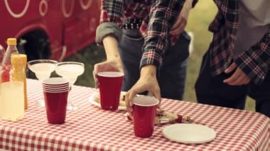 Kareli masa örtüsü, yiyecek ve içeceklerle dolu masaya odaklan. Erkek, kız arkadaşına içki dolduruyor. Barbekü ve açık havada piknik. Parti, arkadaşlık, yaz, boş zaman, aktivite, eğlence ve neşe kavramı