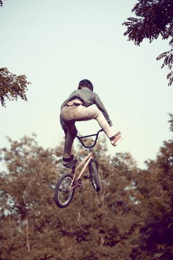 Arka planda, kasklı genç bir adamın ormanda BMX bisiklet sürerken, antrenman yaparken, zor ve tehlikeli numaralar yaparken dinamik görüntüsü. Aktif yaşam tarzı, spor, ekstrem, dinamik, hobi, serbest stil kavramı
