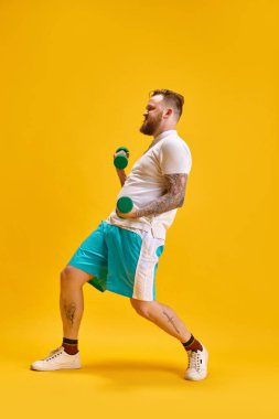 Spor kıyafetleri giymiş sakallı bir adamın sarı arka planda duran dambıllarıyla tam boy görüntüsü. Meme egzersizi. Spor anlayışı, güç, moda, duygular, yaşam tarzı, reklam