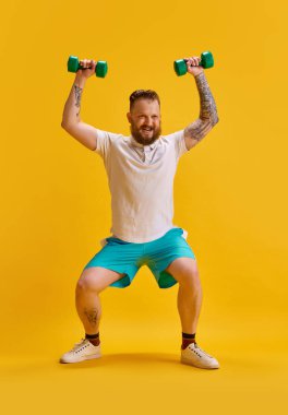 Spor kıyafetleri giymiş sakallı bir adamın sarı arka planda duran dambıllarıyla tam boy görüntüsü. Meme egzersizi. Spor anlayışı, güç, moda, duygular, yaşam tarzı, reklam