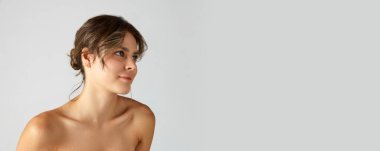 Gri stüdyo arka planında pürüzsüz, sağlıklı, mükemmel bir cilde ve çıplak omuzlara sahip zarif, güzel bir genç kadın. Doğal güzellik kavramı, kozmetik ve dermatoloji, cilt bakımı, kozmetik, reklam. Pankart