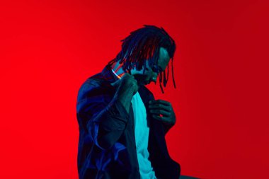 Müzisyen. Kırmızı arka planda, neon ışıkta korkuyla poz veren yakışıklı, şık bir genç. Sanat, moda, modern tarz, siber punk, fütürizm, gençlik kültürü, reklam