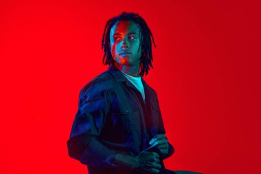 Yüzünde renkli neon filtre yansıması olan yakışıklı Afrikalı genç adam neon ışıkta kırmızı arka planda poz veriyor. Sanat, moda, modern tarz, siber punk, fütürizm, gençlik kültürü, reklam