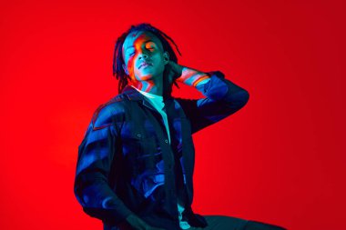 Neon ışıkta kırmızı arkaplana karşı poz veren neon filtre yansımalı genç Afrikalı bir adam. Sürrealizm. Sanat, moda, modern tarz, siber punk, fütürizm, gençlik kültürü, reklam