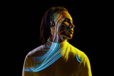 Yüzünde renkli neon çizgileri olan yakışıklı Afrikalı bir adam neon ışıkta siyah arka plana karşı poz veriyor. Sanat, moda, modern tarz, siber punk, fütürizm, reklam