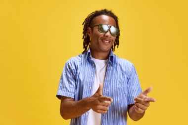 Sarı stüdyo arka planına gülümseyen, kareli tişörtlü ve güneş gözlüklü yakışıklı, şık bir genç. Gençlik kavramı, insan duyguları, yaşam tarzı, moda, yüz ifadeleri, reklam