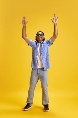 Güneş gözlüklü ve günlük kıyafetlerle sarı stüdyo arka planına rağmen kulaklıkla müzik dinleyen neşeli genç bir adamın tam boy görüntüsü. Gençlik kavramı, duygular, yaşam tarzı, moda, ifade