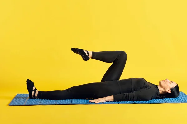 フィットネスマットの上に横たわる黒いスポーツウェアの女性と 黄色いスタジオの背景に伸びる1本の足を上げます スポーツ 健康的なライフスタイル フィットネス ボディケア ウェルネス 広告のコンセプト — ストック写真
