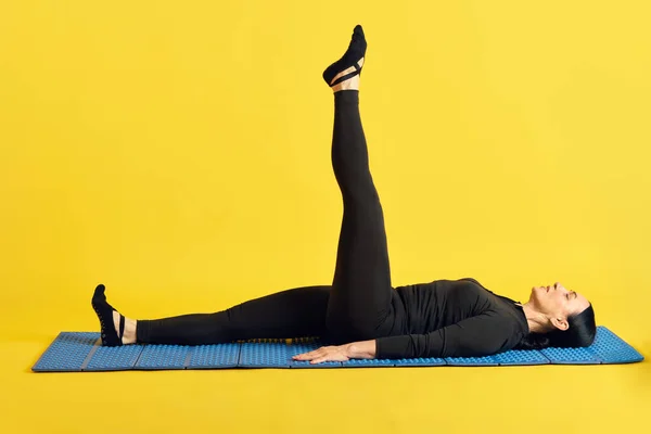 フィットネスマットに横たわる黒いスポーツウェアのスリムな女性と イエロースタジオの背景に伸びた1本の足を上げます スポーツ 健康的なライフスタイル フィットネス ボディケア ウェルネス 広告のコンセプト — ストック写真