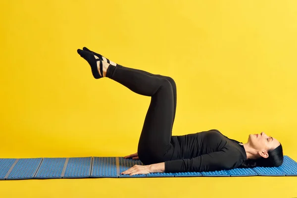 フィットネスマットの上に横たわる黒いスポーツウェアのスポーティブな女性と 黄色いスタジオの背景に伸びる脚 スポーツ 健康的なライフスタイル フィットネス ボディケア ウェルネス 広告のコンセプト — ストック写真