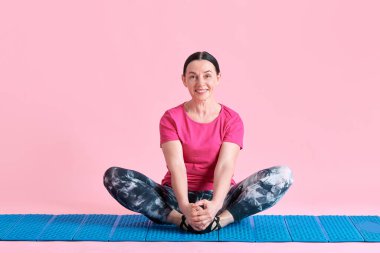 Lotus yoga pozunda spor minderinde oturmuş gülümseyen olgun bir kadın. Pembe stüdyo arka planında meditasyon yapıyor. Spor anlayışı, sağlıklı yaşam tarzı, fitness, vücut bakımı, sağlık, reklam