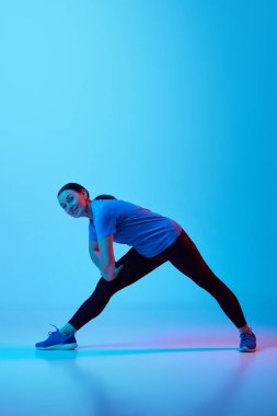 Diz ağrısının azaltılması. Siyah spor giyim giymiş, bacakları esneyen, mavi stüdyo arka planında neon ışıklarıyla antrenman yapan olgun, zayıf bir kadın. Spor anlayışı, sağlıklı yaşam tarzı, fitness, vücut bakımı, sağlık, reklam