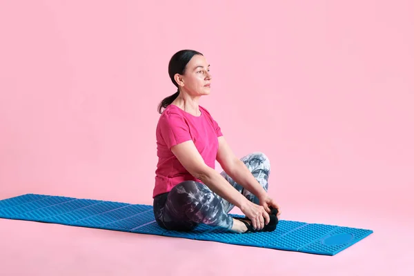 蓮のヨガのポーズでフィットネスマットに座っている美しい成熟した女性 ピンクのスタジオの背景にトレーニングと瞑想 スポーツ 健康的なライフスタイル フィットネス ボディケア ウェルネス 広告のコンセプト — ストック写真