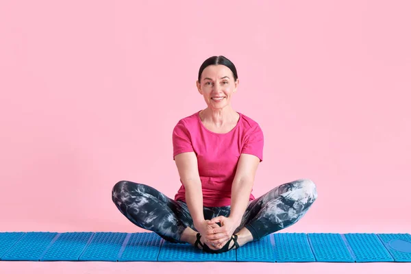笑顔の成熟した女性は 蓮のヨガのポーズでフィットネスマットに座って ピンクのスタジオの背景にトレーニングと瞑想 スポーツ 健康的なライフスタイル フィットネス ボディケア ウェルネス 広告のコンセプト — ストック写真