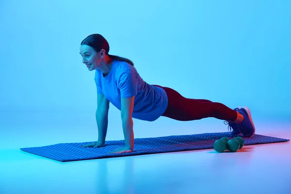 美しい成熟した女性のトレーニングは ネオンライトの青いスタジオの背景に対してプランクに立っています スポーツ 健康的なライフスタイル フィットネス ボディケア ウェルネス 広告のコンセプト — ストック写真