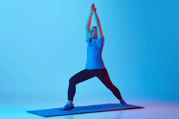 体をトーニングする ネオンライトの青いスタジオの背景に伸びるフィットネスマットの女性トレーニング スポーツ 健康的なライフスタイル フィットネス ボディケア ウェルネス 広告のコンセプト — ストック写真
