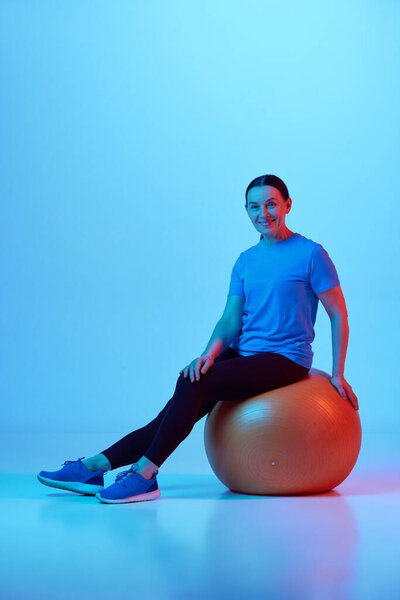 Улыбающаяся взрослая женщина, сидящая на фитнес-мяче после тренировки на синем фоне студии в неоновом свете. Концепция спорта, здоровый образ жизни, фитнес, уход за телом, оздоровление, реклама