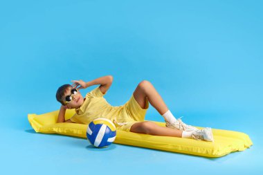 Keyif. Mutlu ve rahat küçük çocuk, yüzme yatağında uzanan çocuk voleybol ile mavi stüdyo arka planına karşı. Çocukluk kavramı, çocuk duyguları, tatil, yaz, eğlence ve neşe, reklam