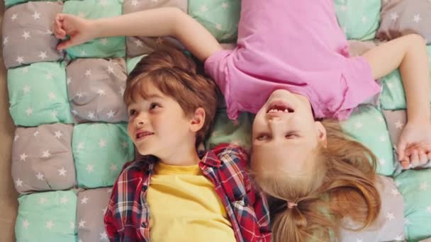 快乐的小男孩和女孩们 兄弟姐妹们躺在地板上 躺在五彩缤纷的毯子上 开心地聊天 爱和照顾的概念 — 图库视频影像