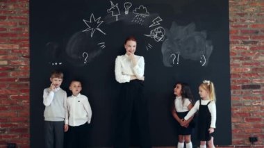 Resmi giyinmiş genç, neşeli bir kadın karatahtanın yanında durup ikonlar çiziyor ve çocuklarla oynuyor. Eğitim ve oyun. İş konsepti, eğitim, dersler, oyunlar, öğrenim, psikoloji, reklam