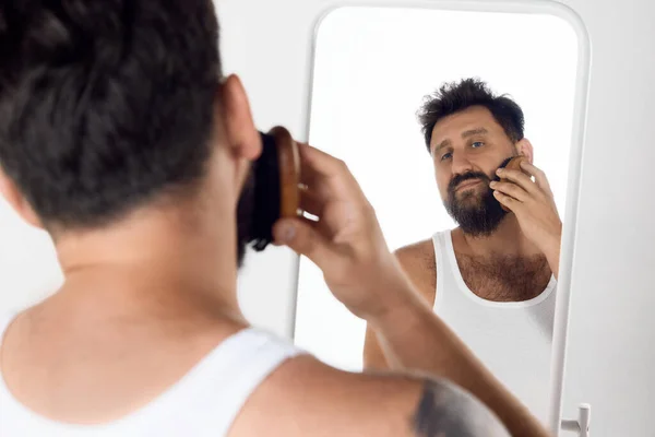 白いスタジオの背景に特別なブラシでブラシをかけ ひげの世話をするミラーを見ている成熟した男 メンズビューティー スキンケア 化粧品 ウェルネスのコンセプト Adのためのスペースをコピーする — ストック写真