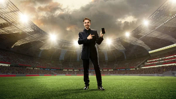 Business in a suit standing on 3d sports arena with flashlights and pointing on mobile phone screen. Betting before game. Concept of sport, fan, betting and finances, gambling, bookmaker