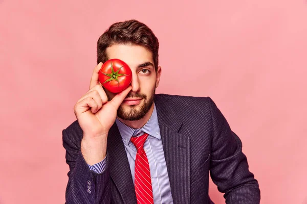 古典的なスーツと赤いネクタイの男は 目の近くにトマトを保持し ピンクのピンクの背景に対してカメラを見ています 食べ物 創造性 健康について ポップアート写真 Adのためのスペースをコピーする — ストック写真