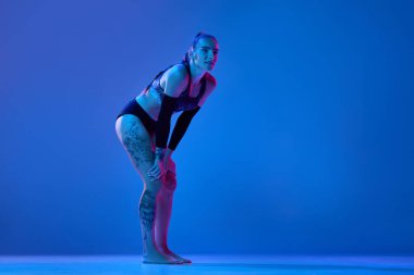 Kadın, spor giyim sektöründe profesyonel bir atlet ellerini dizlerinin üstüne çöküp neon ışıkta mavi stüdyonun arka planına yaslanıyor. Profesyonel spor kavramı, rekabet, sağlık, hobi, eylem, reklam