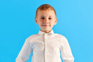 Küçük bir çocuğun portresi, ilkokul öğrencisi, beyaz tişörtlü bir çocuk mavi stüdyo arka planında gülümseyerek kameraya bakıyor. Çocukluk kavramı, eğitim, okul, çocuk duyguları, reklam