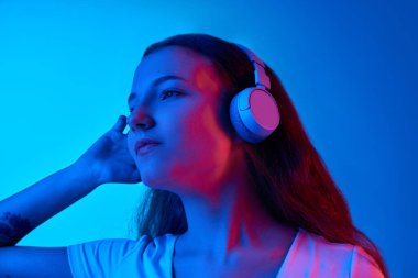 Kulaklıkla müzik dinleyen bir kız mavi stüdyonun arka planına karşı neon ışıkta. Keyif ve rahatlama. İnsan duyguları kavramı, moda, güzellik, yaşam tarzı, gençlik, reklam