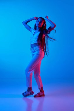 Gündelik giysiler içinde kulaklıkla müzik dinleyen ve neon ışıkta mavi stüdyonun arka planında dans eden genç bir kız. İnsan duyguları kavramı, moda, güzellik, yaşam tarzı, gençlik, reklam