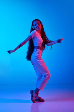 Gündelik giysiler içinde kulaklıkla müzik dinleyen ve neon ışıkta mavi stüdyonun arka planında dans eden genç bir kız. İnsan duyguları kavramı, moda, güzellik, yaşam tarzı, gençlik, reklam
