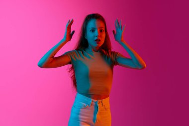 Genç duygusal kız ellerini kaldırıyor, geniş açık gözlerle, pembe stüdyonun arka planına karşı şok olmuş bir şekilde neon ışıkta duruyor. İnsan duyguları kavramı, moda, güzellik, yaşam tarzı, gençlik, reklam