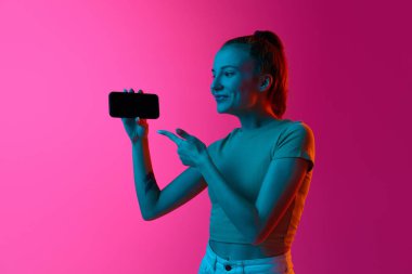 Gülümseyen genç kız cep telefonu ekranını işaret ediyor. Pembe stüdyo arka planında, neon ışıkta. Satış, çevrimiçi hizmetler. İnsan duyguları kavramı, sosyal medya, satış, alışveriş, gençlik, reklam