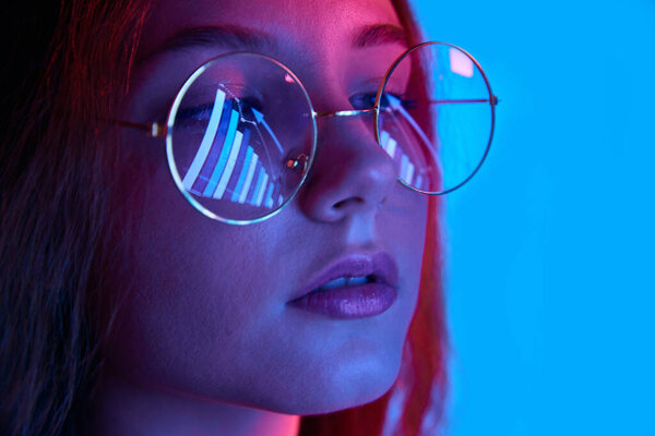 Close-up of female face in glasses with analyticals graph reflection against blue studio background in neon light. Concept of human emotions, business, trade market, lifestyle, youth, ad