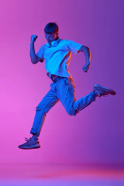 stock image Full-length image of young guy in casual clothes and headphones jumping over gradient pink purple background in neon light. Concept of human emotions, youth, lifestyle, fashion, facial expressions, ad