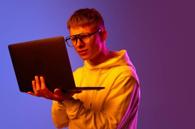 Genç bir adam, öğrenci, gözlüklü serbest çalışan dikkatle dizüstü bilgisayara bakıyor ve neon ışıkta mor bir arka plan görüyor. İnsan duyguları kavramı, gençlik, yaşam tarzı, iş, eğitim, reklam